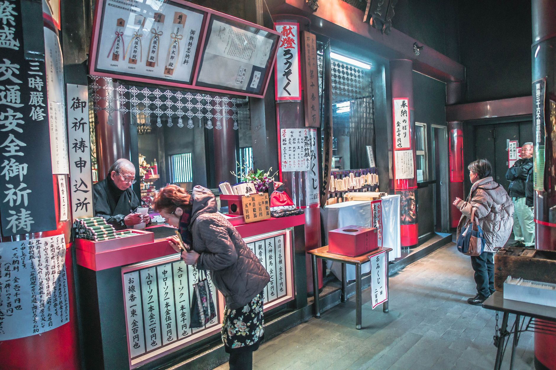 Guichet d'un temple japonais tenu par un religieux, avec tarifs de bougies et d'encens, demandes de prières et boîte à omikuji