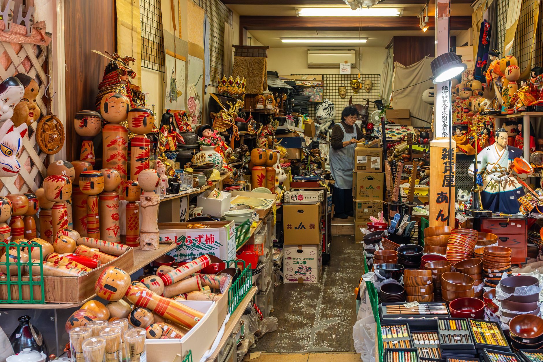 Étals d'une boutique d'objets japonais : dizaines de poupées kokeshi en bois à tête ronde et corps cylindrique peints de motifs rouges, à côté de bols en bois et de pinceaux