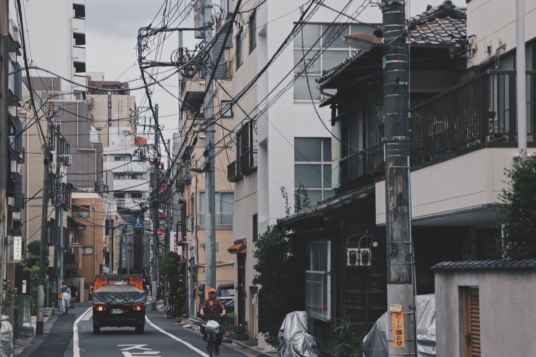 Rue résidentielle étroite au Japon, bordée de poteaux électriques et de petits immeubles, avec un camion et un cycliste au loin