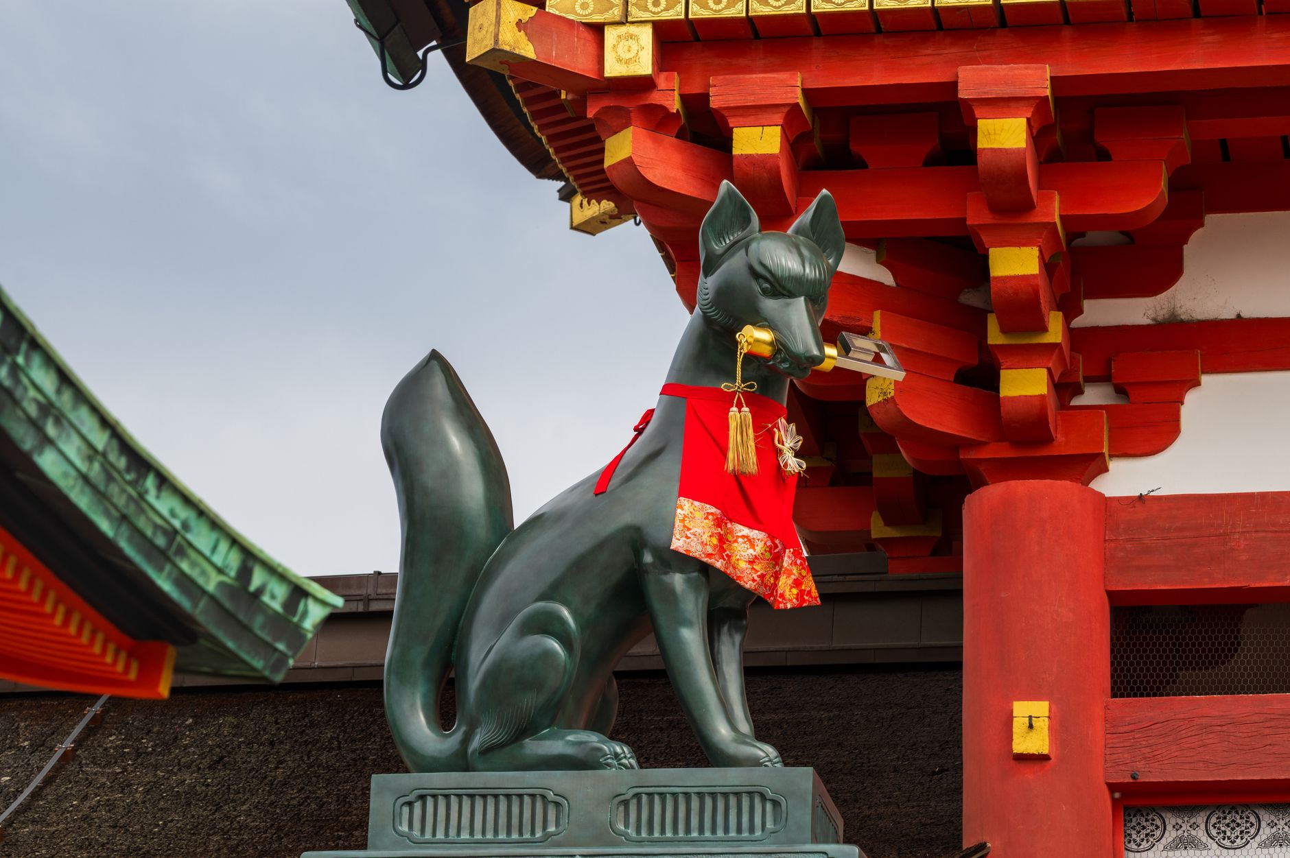 Statue de renard en bronze vert portant un bavoir rouge, un objet doré dans la gueule, devant un bâtiment de sanctuaire vermillon