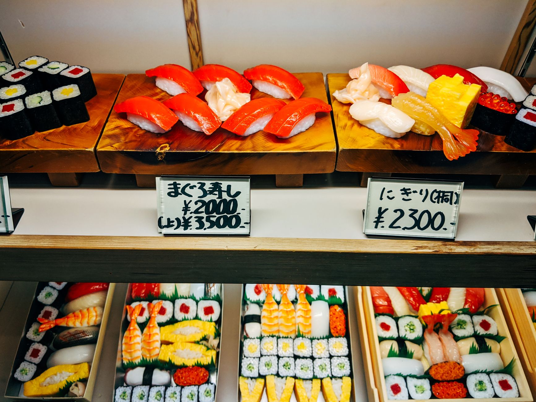 Vitrine de restaurant japonais présentant des modèles de sushis et de makis avec des étiquettes de prix manuscrites en yens