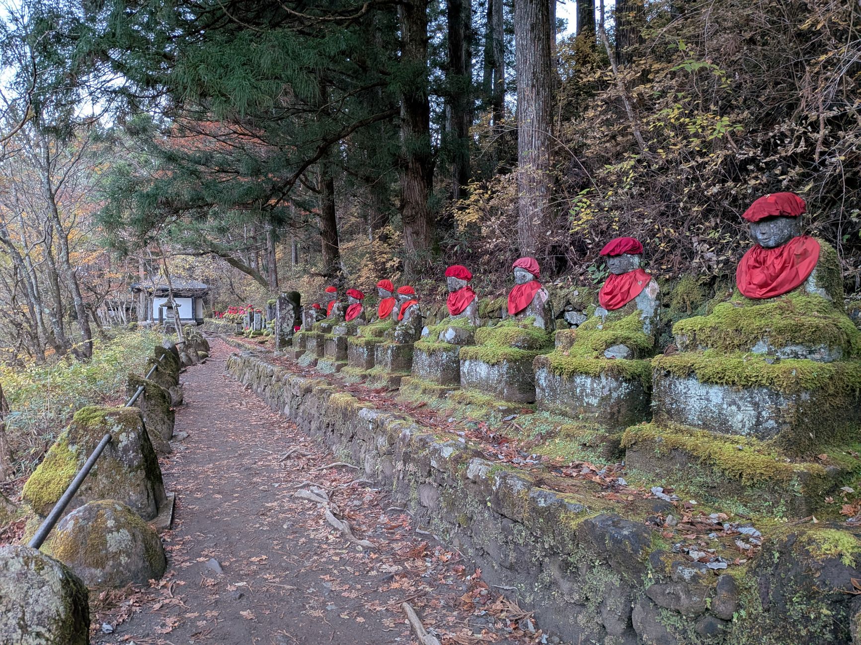 Rangée de statues de jizō en pierre couvertes de mousse, coiffées de bonnets rouges et portant des bavoirs rouges, le long d'un chemin forestier en automne