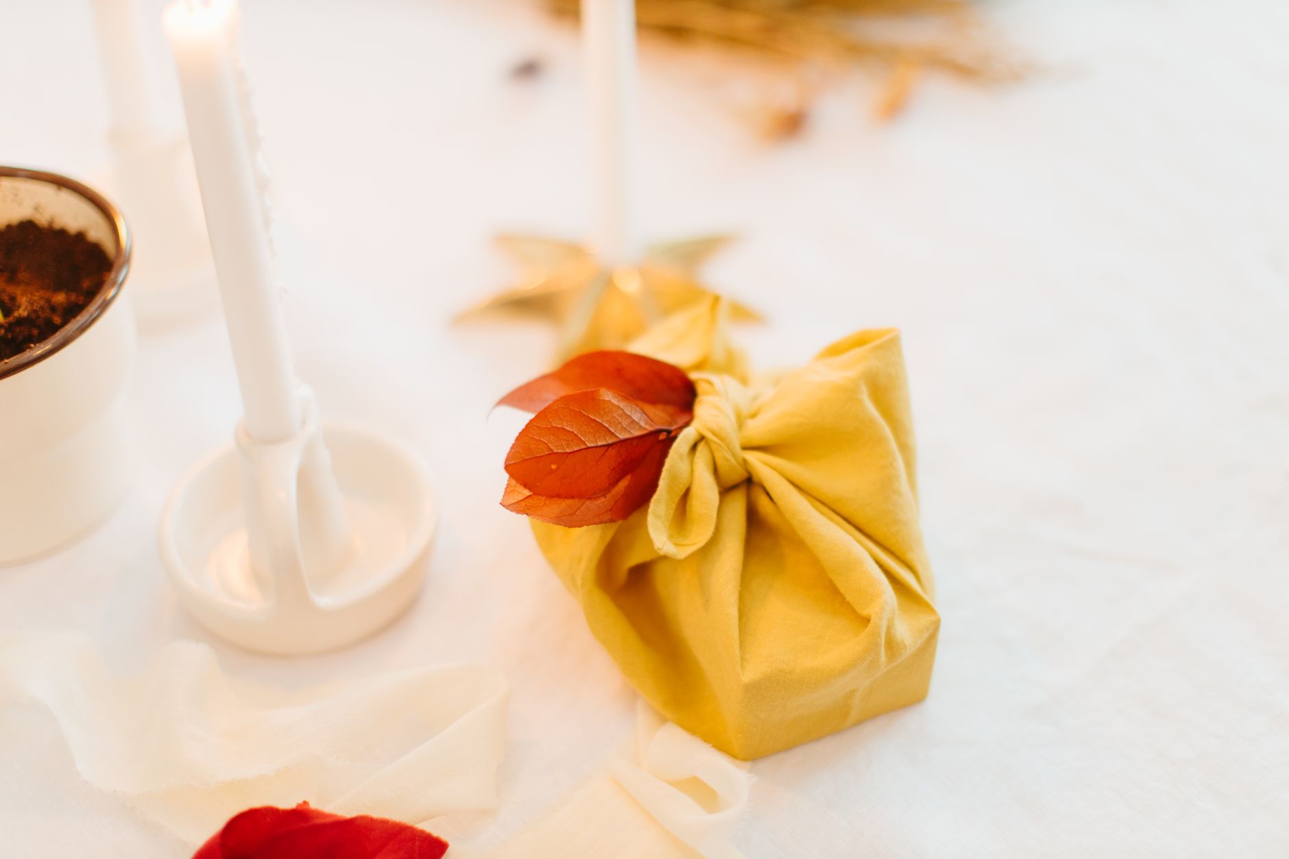 Petit paquet enveloppé dans un tissu jaune noué sur le dessus, orné d'une feuille rousse, posé sur une table près de bougies
